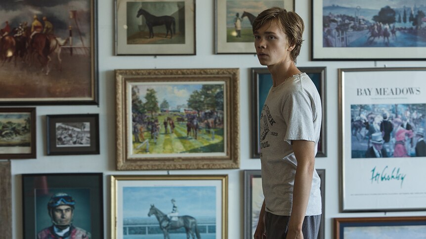 Colour still of Charlie Plummer standing in front of wall of horse artworks in 2018 film Lean on Pete.