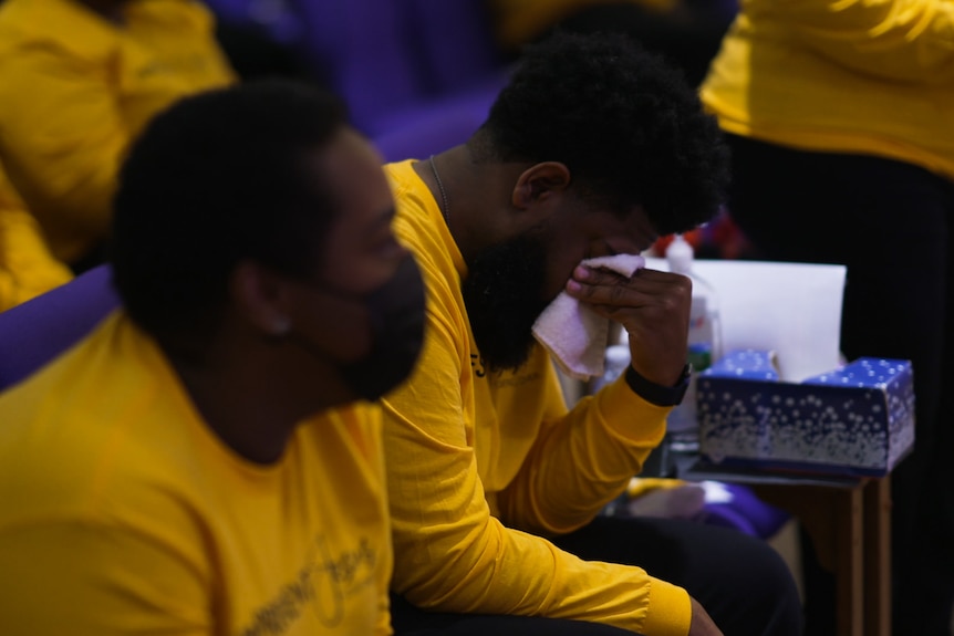 A man leans over his lap and cries into his tissue while sitting with others in church pews.