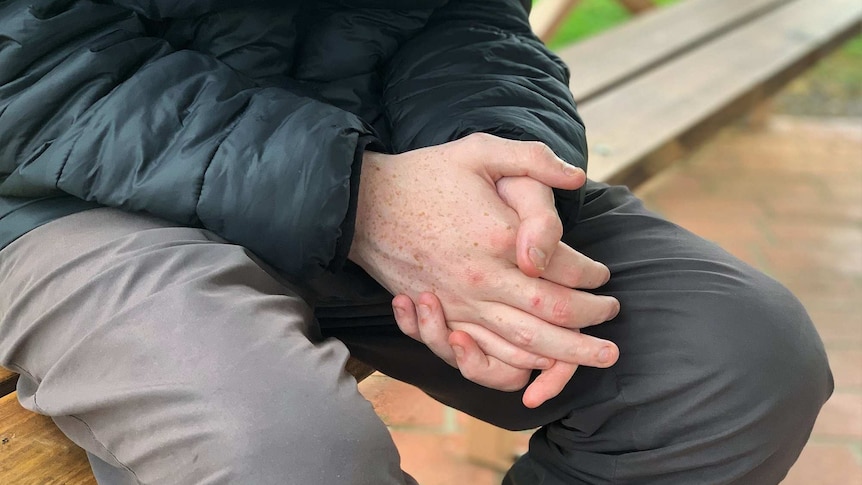 A teenage boy sits on a park bench with his hands clasped together.