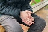 A teenage boy sits on a park bench with his hands clasped together.