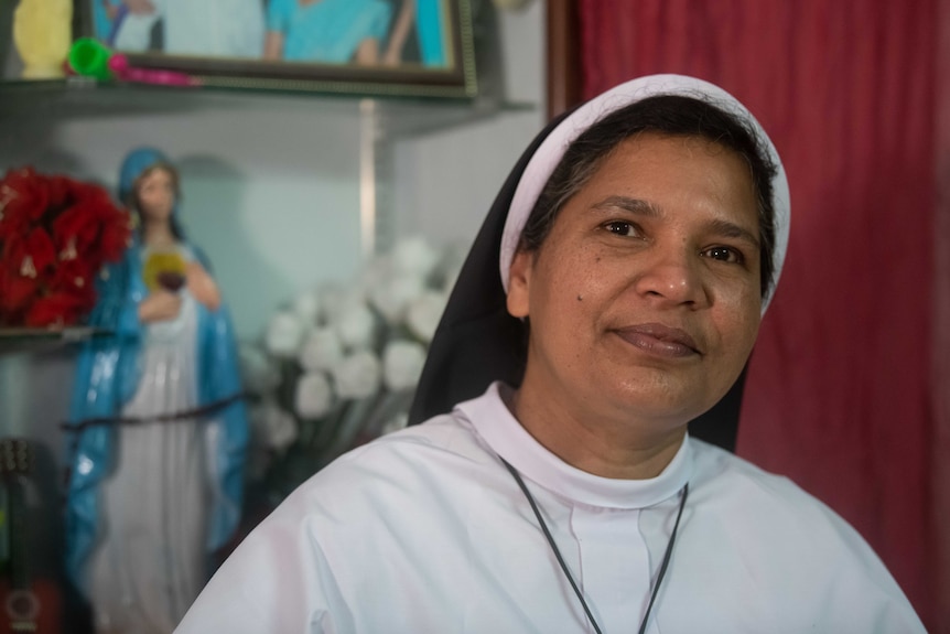 A nun in a habit looks at the camera, there's a statue of the Virgin Mary behind her.