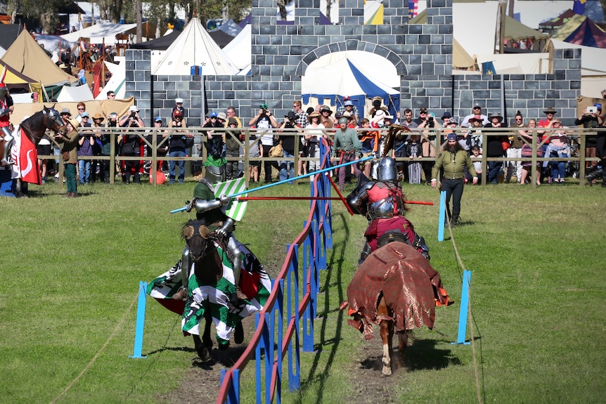 Two riders on horseback brake lances against each other's shields as the crowd looks on.