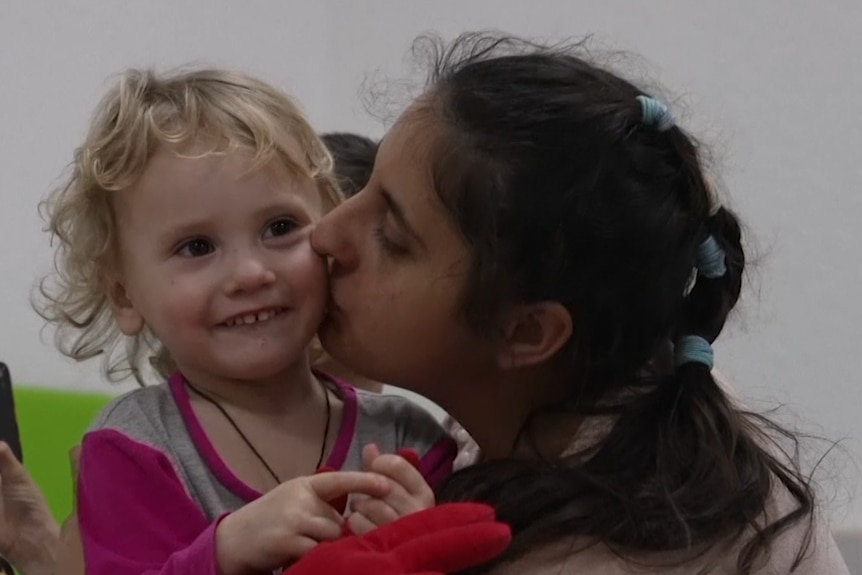 A brown-haired woman plants a kiss on a smiling blonde toddler's cheek