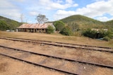 Rusting old building with veranda stands beside rail lines, beneath low hills 