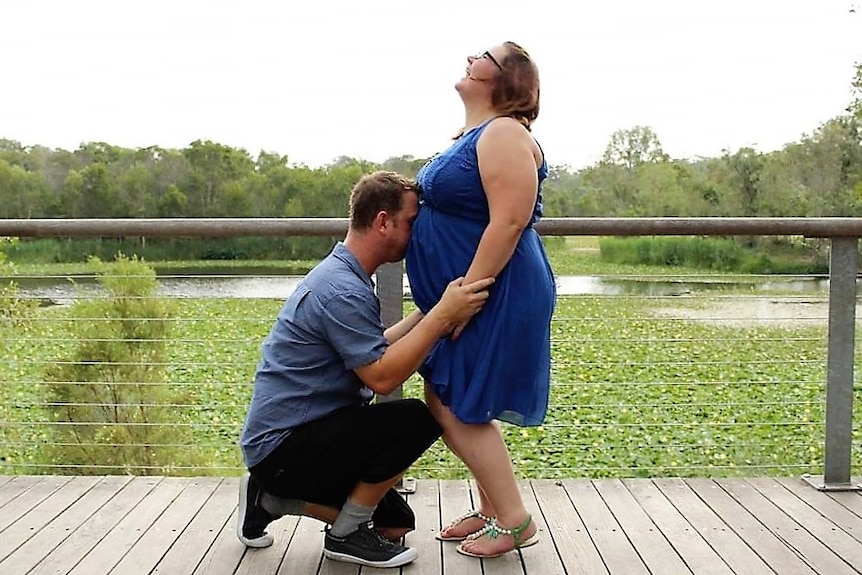 A pregnant lady in a blue dress laughing, as her husband kisses her belly.