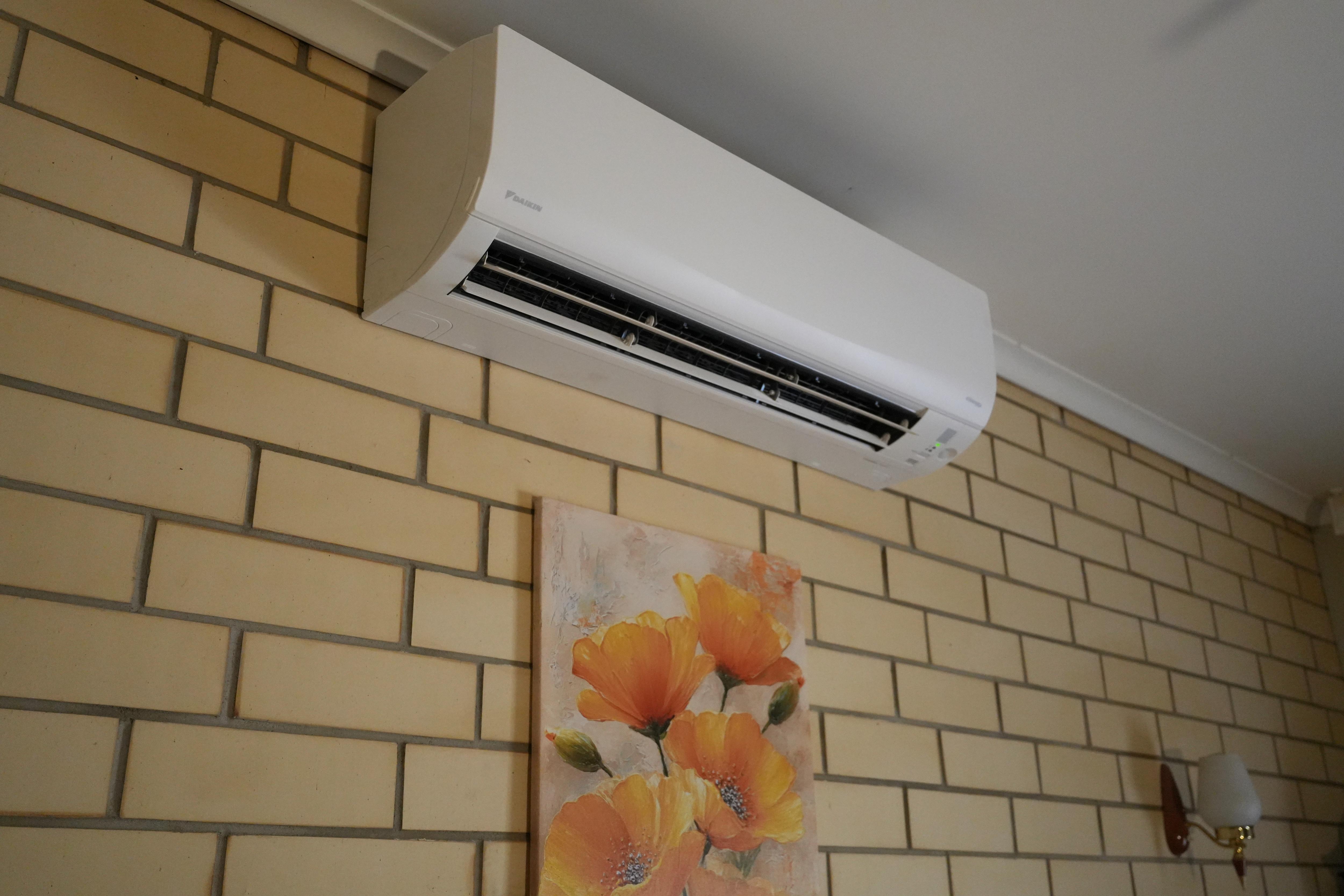 Um ar condicionado branco está pendurado em uma parede de tijolos bege. Abaixo está a imagem de uma flor de laranjeira.