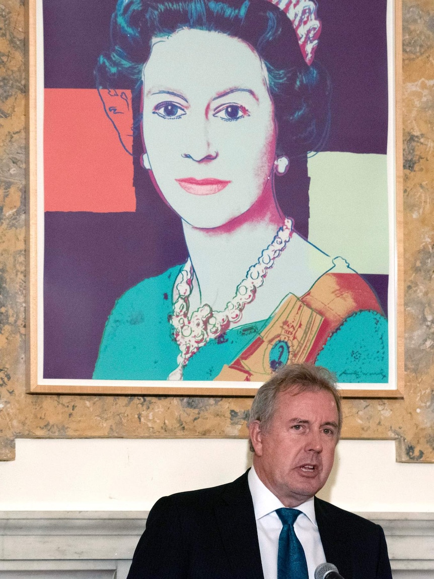 A large modernist painting of Queen Elizabeth II hangs over Sir Kim Darroch as he speaks in front of a sand marble wall.