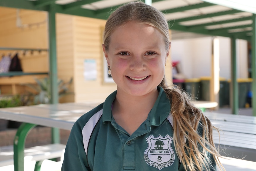 A young girl wearing a green school shirt and blonde hair tied back smiling in the school yard.