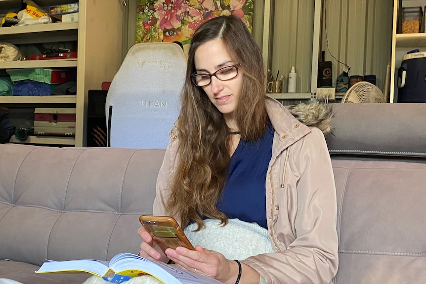 A pregnant woman with long, dark hair sits on a couch looking at her phone.