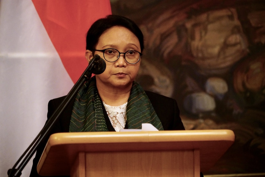 A woman with glasses stands at a lectern looking stern