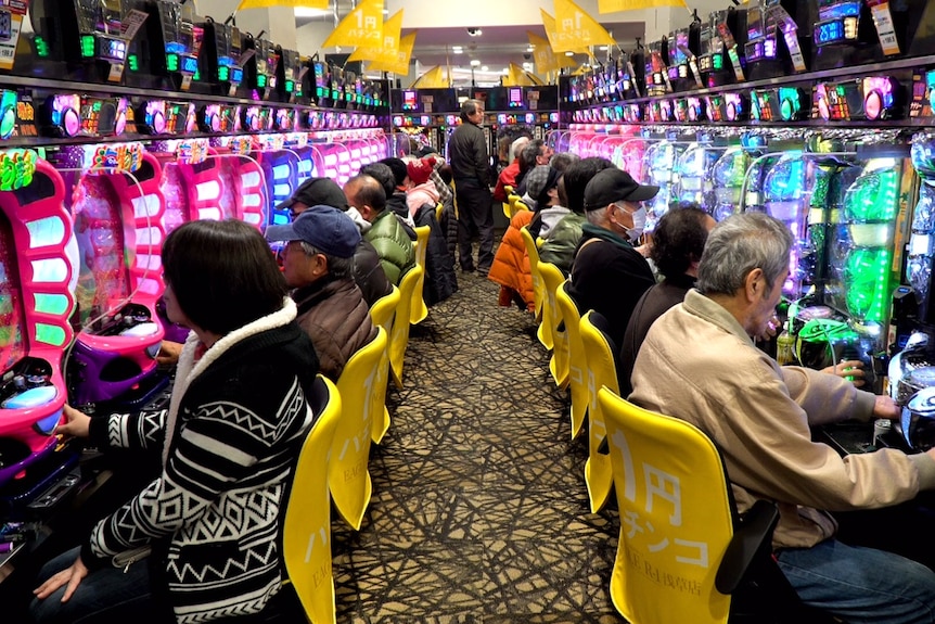 Rows of people inside a venue on brightly-lit machines playing Pachinko.