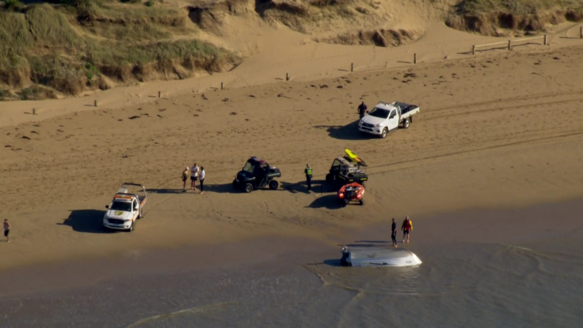Emergency vehicles are parked on the beach and people, including lifesavers and police, gather around an overturned boat.