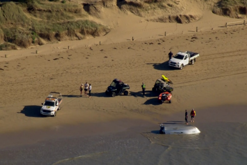 Emergency vehicles are parked on the beach and people, including lifesavers and police, gather around an overturned boat.
