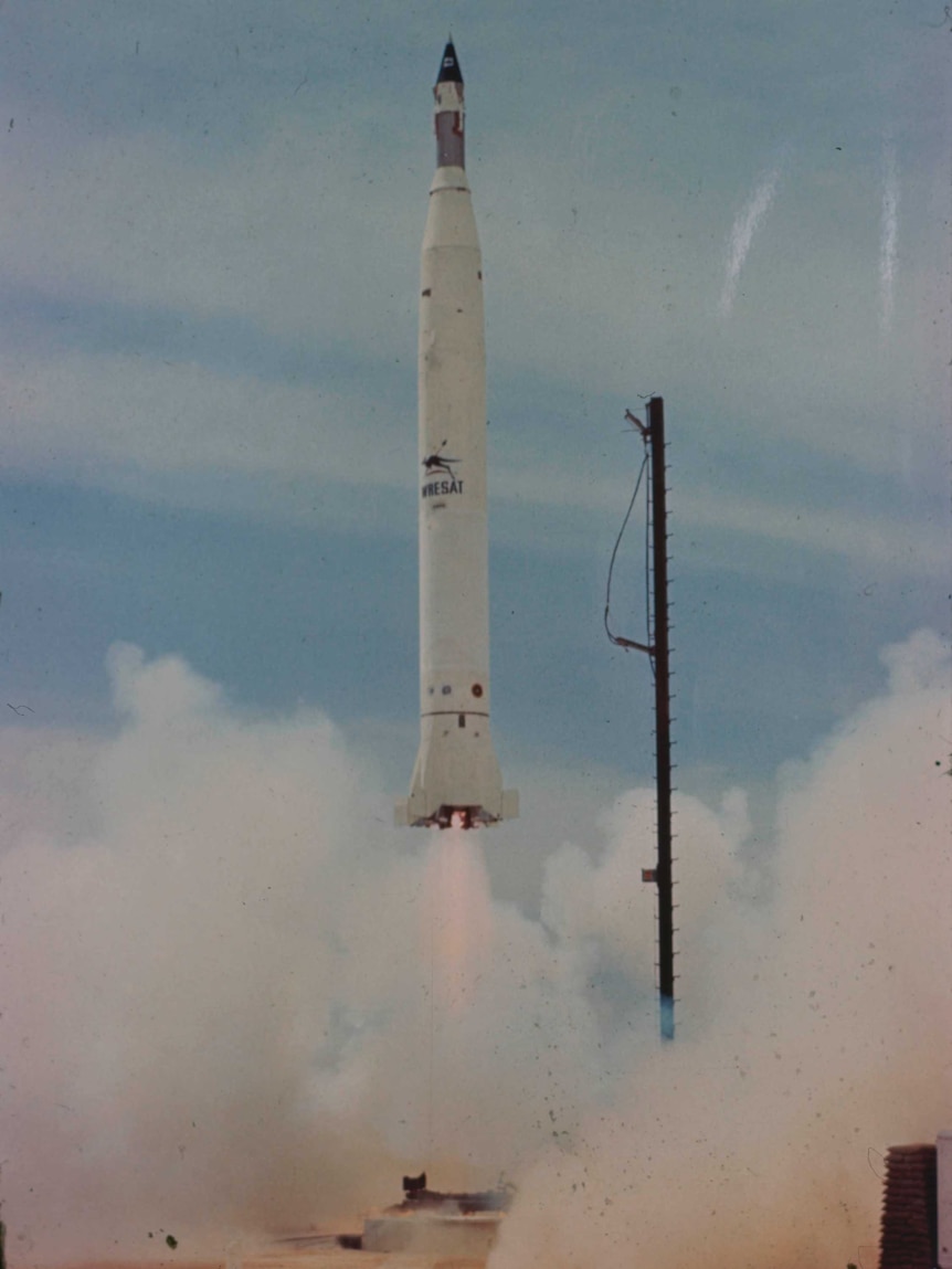 An old coloured photo shows a rocket being launched over outback red dirt against a blue sky.