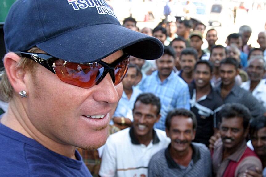 Shane Warne, wearing a blue cap and wraparound sunglasses, stands in front of a crowd of smiling fans