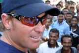 Shane Warne, wearing a blue cap and wraparound sunglasses, stands in front of a crowd of smiling fans