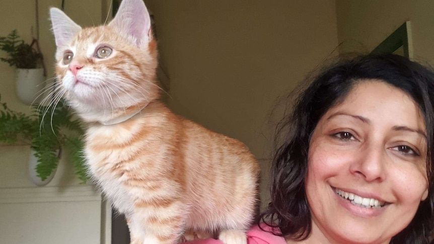 A woman stands with a ginger kitten on her shoulder.