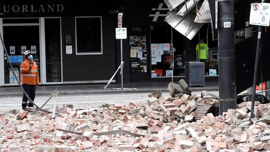 A person in high vis looks at building with bricks scattered on the ground.