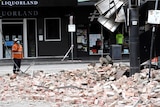 A person in high vis looks at building with bricks scattered on the ground.