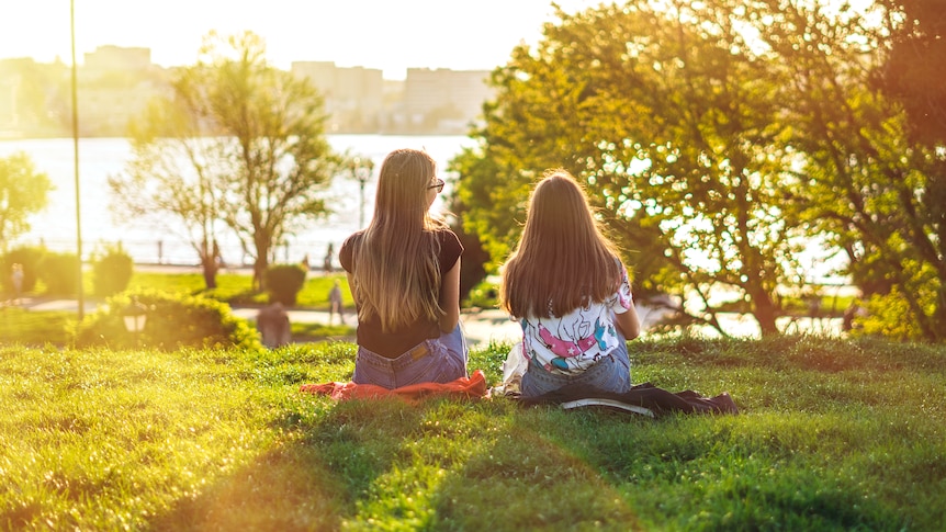 Two young women sitting on the grass in a park basked in golden afternoon light as they look out over water. 