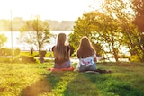 Two young women sitting on the grass in a park basked in golden afternoon light as they look out over water. 