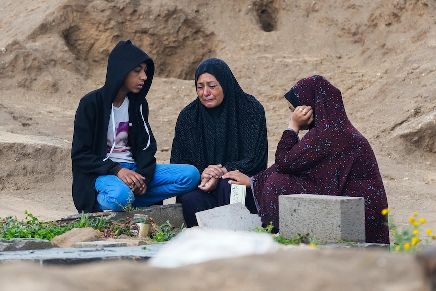 A woman kneels next to a grave crying alongside two other people who are consoling her