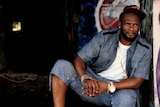 DJ Just Dizle, a black man wearing denim shirt and shorts with a white shirt underneath, looking away from camera with cap