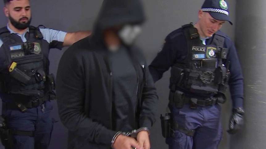 Two police lead a man wearing a hood and mask