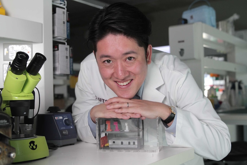 Dr Joshua Chou resting his hands and head on a box-shaped prototype of the device that will take the cancer cells into space
