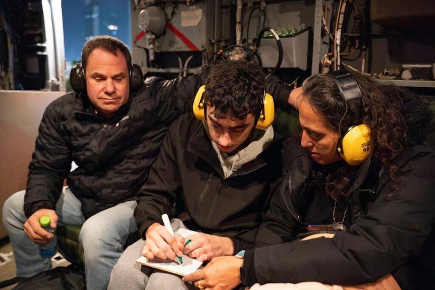 A teenager sits between two adults, all wearing ear muffs, sitting inside a helicopter. He writes on a notepad
