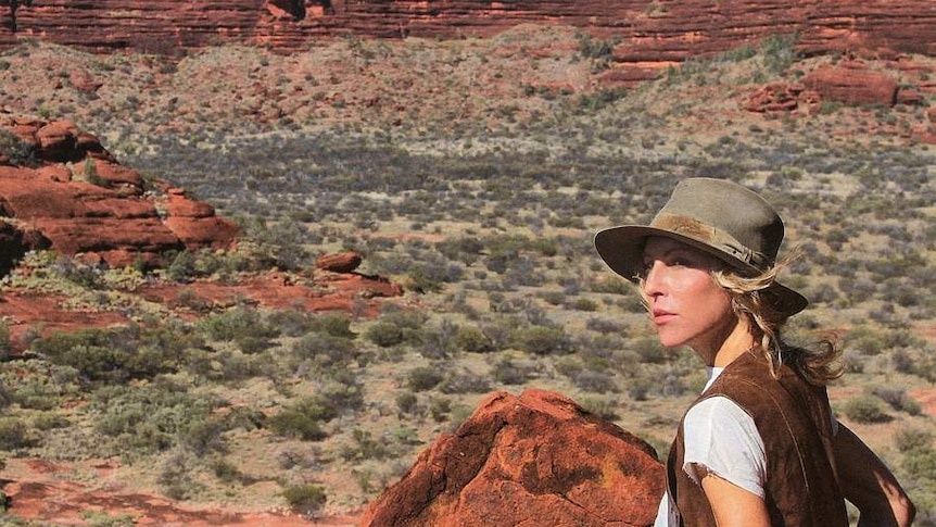 A woman looks out at a red dessert landscape, marked with small shurbs and rocky hills.