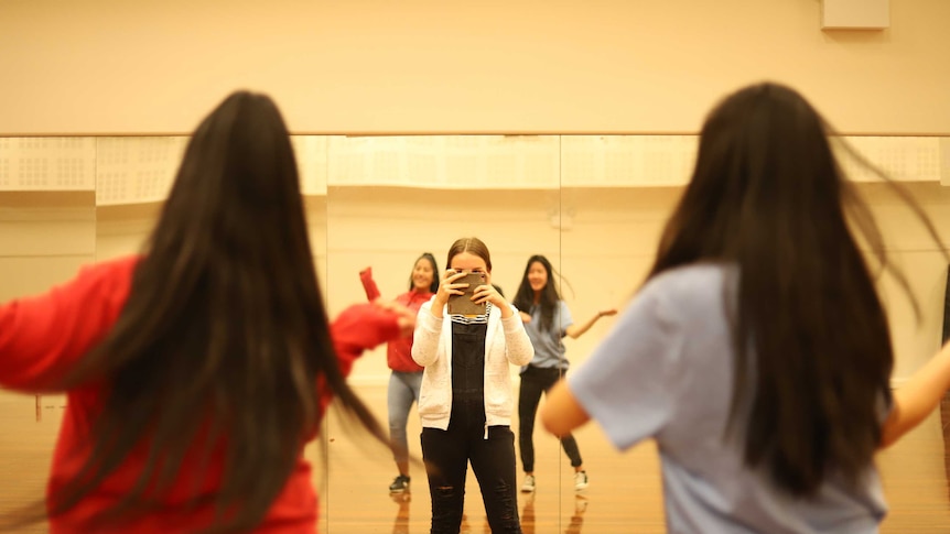 Two girls dance in front of mirror in a dance studio, while facing a third girl who is recording the routine on her mobile phone