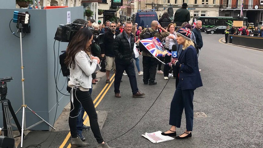 A woman in a suit with a union jack beanie and gloves holds a microphone as she speaks to a camera on a packed street