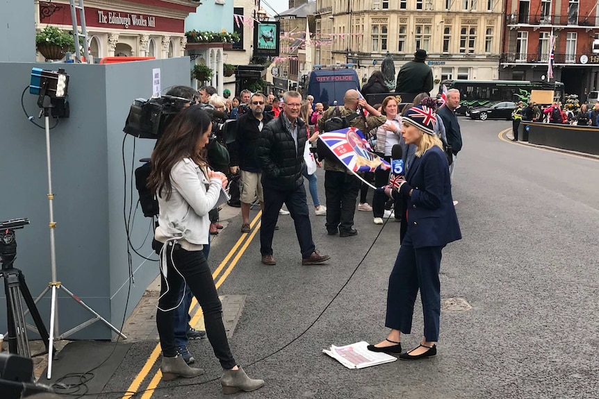 A woman in a suit with a union jack beanie and gloves holds a microphone as she speaks to a camera on a packed street