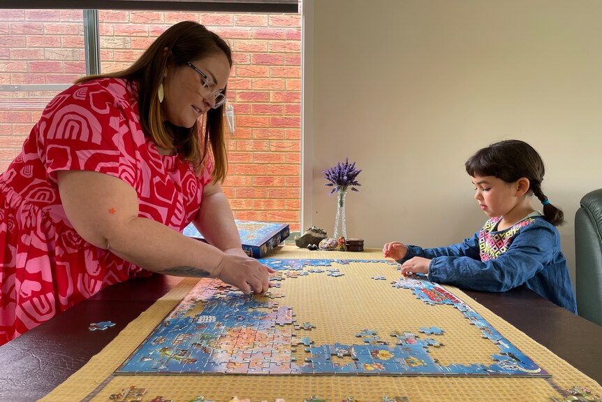 a woman doing a puzzle on a table with her young daughter. There is a vase of purple flowers behind them.
