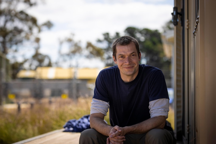 A man sits with his elbows resting on his thighs and hands clasped