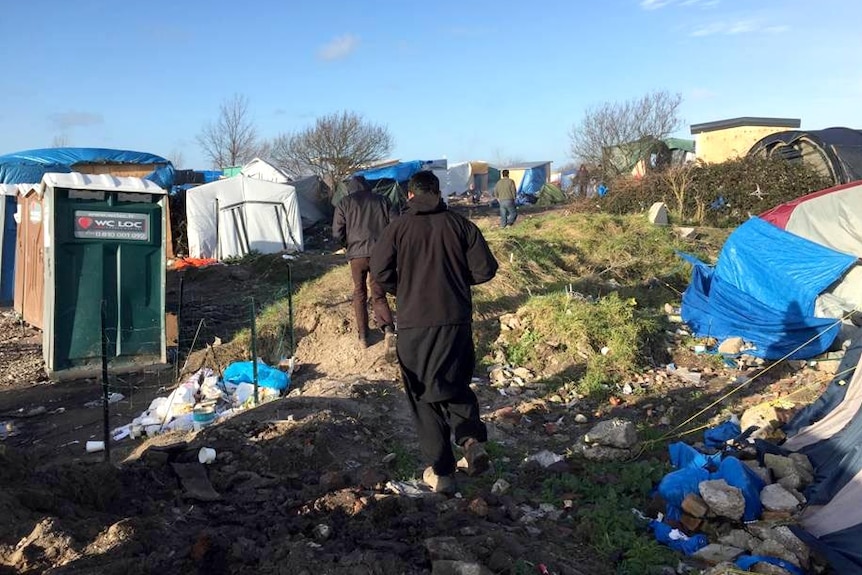 Refugees walk through the refugee camp, surrounded by tents and rubbish on the ground.