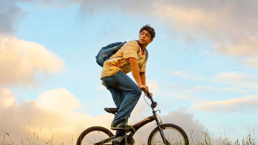 A teenage boy with a backpack and baggy jeans, riding a bike through the grass, looks behind him 