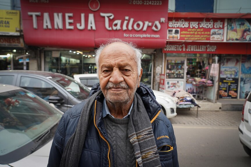 An older man stands on a street with cars passing by behind and a large red sign reading Taneja Tailors