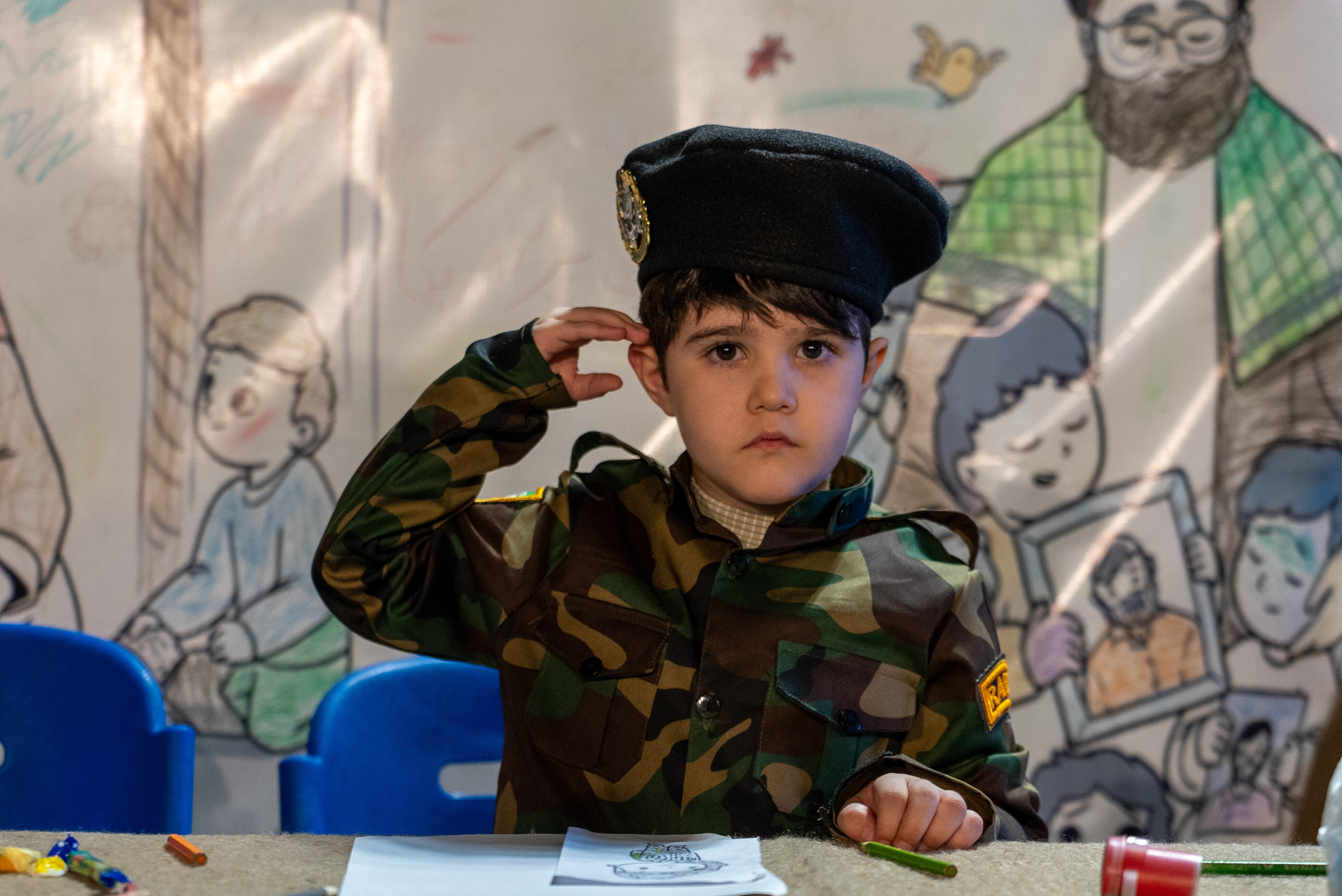 A young boy in a military uniform sitting at a chair with paper and crayons in front of him. He does a salute.