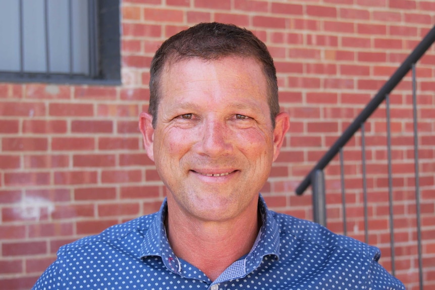 A man with short hair stands smiling in front of a wall of bricks.