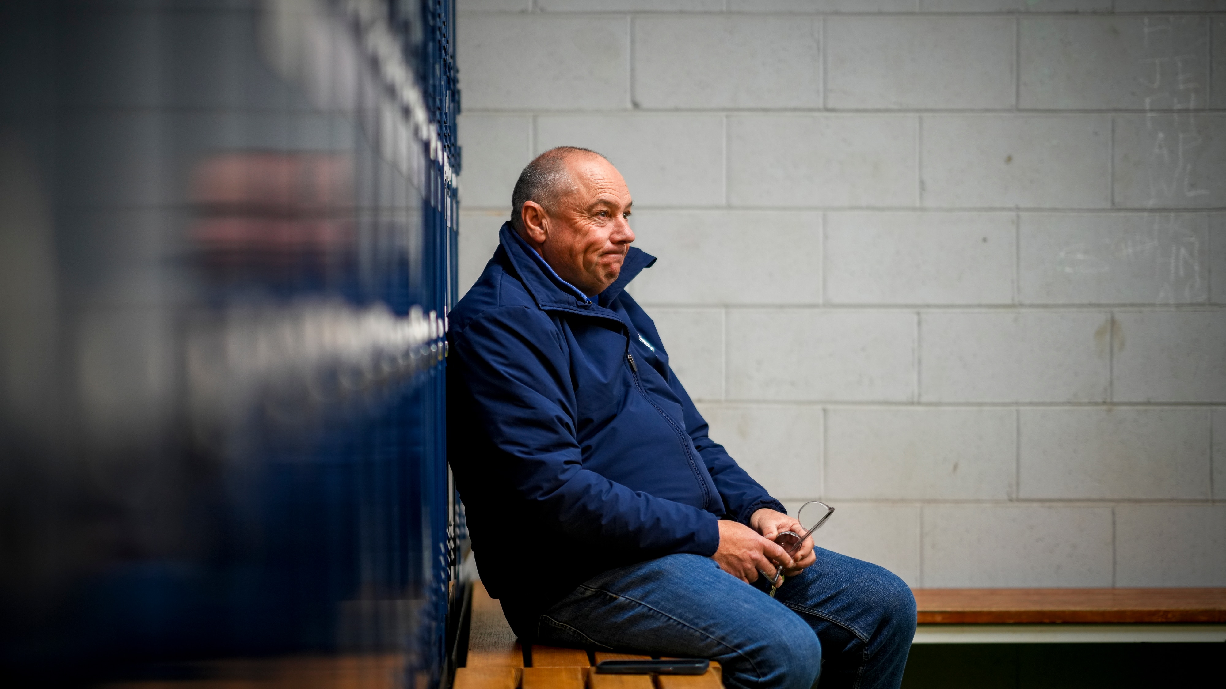 A man looking pensive sits on a bench in a locker room.
