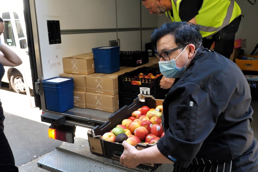 A woman wearing a blue mask and black clothes carries a box of apples from a truck