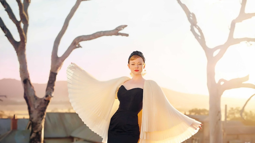 A woman in formal wear stands on a rock above a country town, flanked by dead trees.