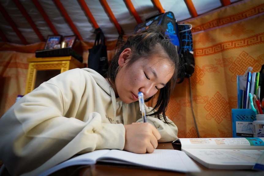 A teenage girl in cream hooodie sits at a desk writing on an open exercise book with a biro