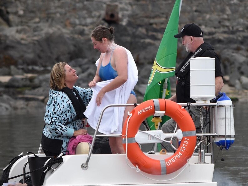 A swimmer stands on a boat with a towel wrapped around her as a person kneels talking to her