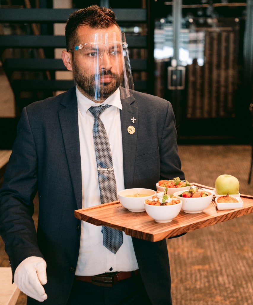 A waiter wears gloves and a face shield while serving a tray of food.