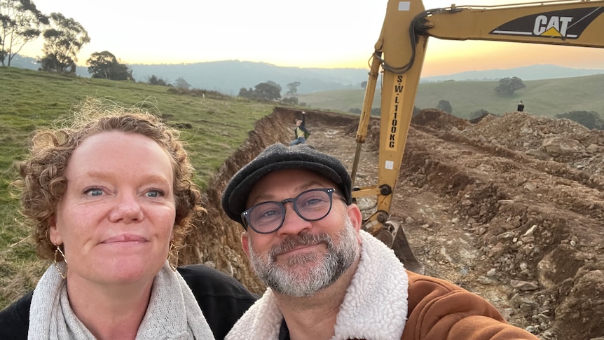 Mandy McCracken and her husband pose smiling for a selfie in front of empty land and an excavator