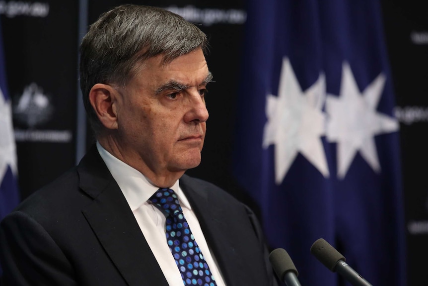 Brendan Murphy stands in front of two microphones. There are Australian flags behind him.