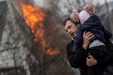 A man runs with a child in his arms, away from a building on fire.